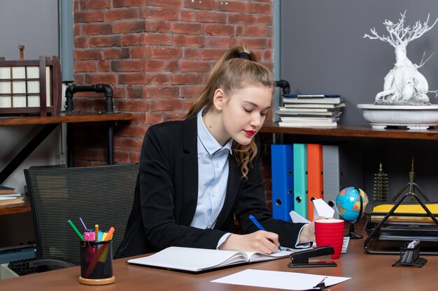 Vue de face d'une jeune femme concentrée assise à une table et écrivant sur un document au bureau