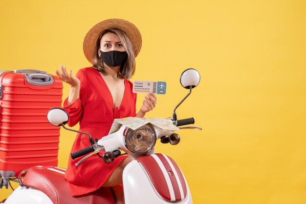 Vue de face d'une jeune femme confuse sur un cyclomoteur avec valise tenant un billet