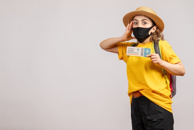 Vue de face de la jeune femme confuse avec masque noir tenant un billet de voyage sur un mur blanc