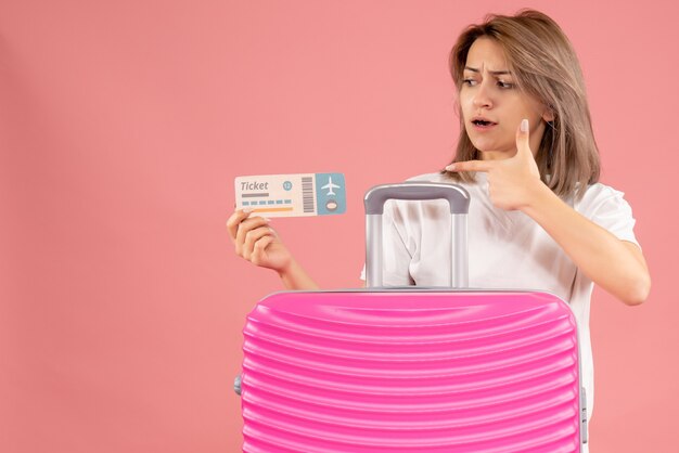 Vue de face de la jeune femme confuse, pointant sur le billet derrière la valise rose