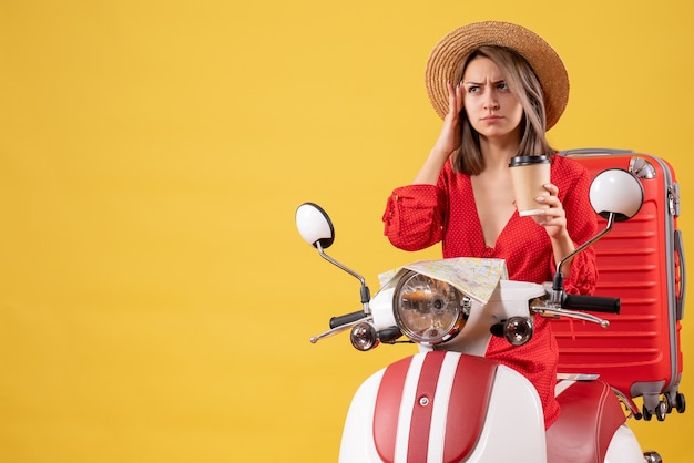 Vue de face de la jeune femme confuse en robe rouge tenant une tasse de café près de cyclomoteur