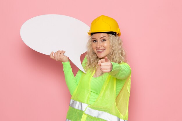 Vue de face jeune femme constructeur en costume de construction vert casque jaune tenant une pancarte blanche avec sourire sur le travail de construction d'architecture d'emploi de l'espace rose