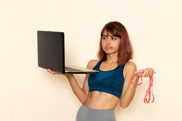 Vue de face de la jeune femme avec corps en forme en chemise bleue à l'aide d'un ordinateur portable sur le mur blanc