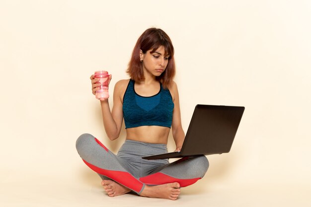 Vue de face de la jeune femme avec un corps en forme de chemise bleue à l'aide d'un ordinateur portable sur un mur blanc