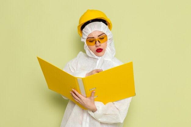 Vue de face jeune femme en costume spécial blanc et casque jaune tenant des fichiers jaunes et écrivant sur la science uniforme du costume d'espace vert