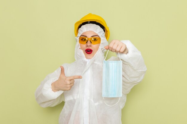 Vue de face jeune femme en costume spécial blanc et casque jaune tenant un masque sur l'espace vert