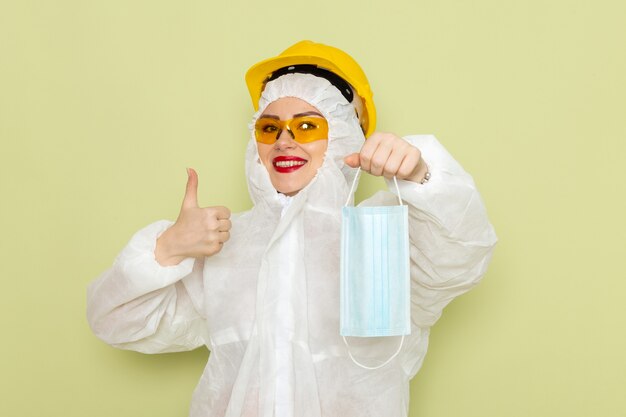 Vue de face jeune femme en costume spécial blanc et casque jaune tenant un masque stérile avec un léger sourire sur le travail de chimie de l'espace vert s