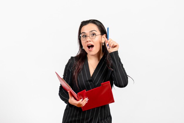 Vue de face jeune femme en costume strict sombre tenant un fichier rouge avec un stylo sur fond blanc bureau d'affaires mode d'emploi document féminin