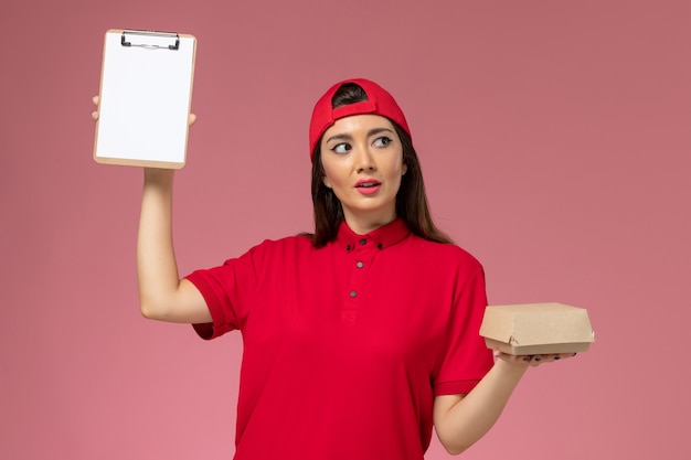 Vue de face jeune femme courrier en cape uniforme rouge avec peu de colis de nourriture de livraison et bloc-notes sur ses mains sur le mur rose