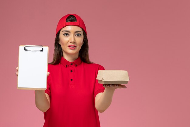 Vue de face jeune femme courrier en cape uniforme rouge avec peu de colis de nourriture de livraison et bloc-notes sur ses mains sur le mur rose