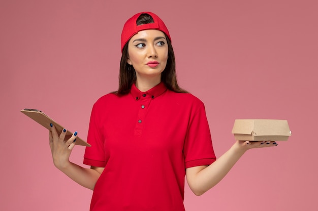 Vue de face jeune femme courrier en cape uniforme rouge avec peu de colis de nourriture de livraison et bloc-notes sur ses mains sur le mur rose