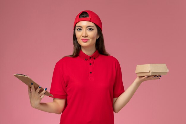 Vue de face jeune femme courrier en cape uniforme rouge avec peu de colis de nourriture de livraison et bloc-notes sur ses mains sur le mur rose