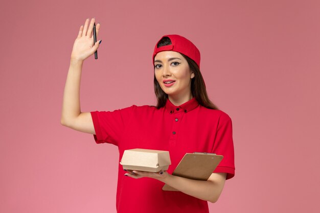 Vue de face jeune femme courrier en cape uniforme rouge avec peu de colis de nourriture de livraison et bloc-notes avec stylo sur ses mains sur le mur rose clair