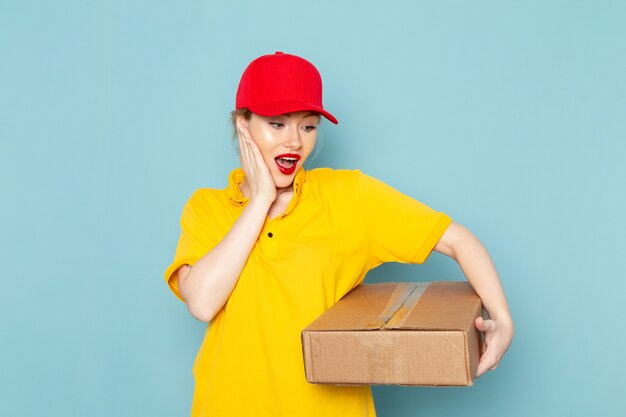 Vue de face jeune femme courrier en chemise jaune et cape rouge souriant et tenant le paquet sur l'espace bleu