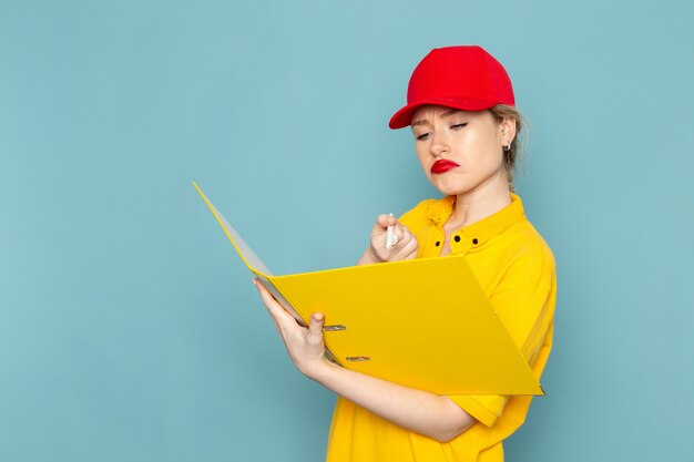 Vue de face jeune femme courrier en chemise jaune et cape rouge tenant un fichier jaune en écrivant des notes sur l'emploi de travailleur de l'espace bleu