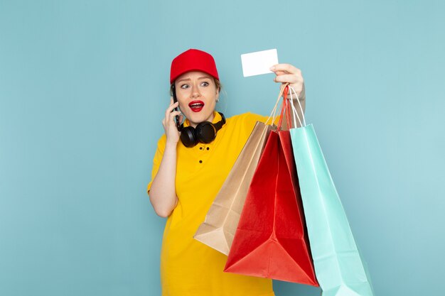 Vue de face jeune femme courrier en chemise jaune et cape rouge tenant des paquets d'achat téléphone et carte sur le travail de plancher bleu