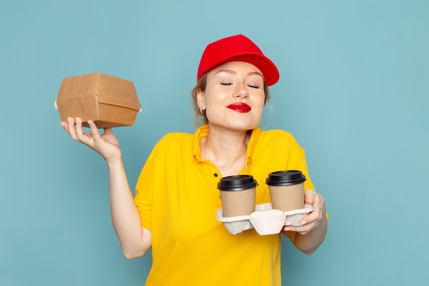 Vue de face jeune femme courrier en chemise jaune et cape rouge tenant des tasses à café en plastique et emballage alimentaire sur l'espace bleu