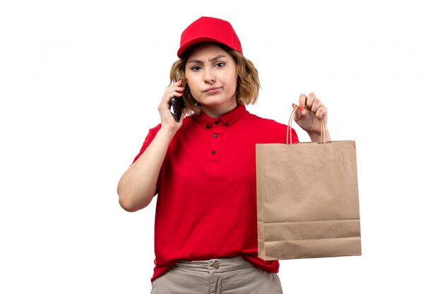 Une vue de face jeune femme courrier en chemise rouge bonnet rouge tenant le colis de livraison parler au téléphone