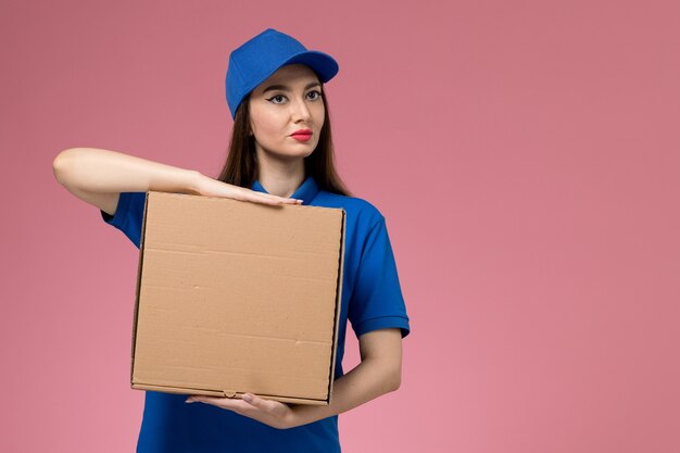 Vue de face jeune femme courrier en uniforme bleu et cape tenant la boîte de livraison de nourriture sur un mur rose clair travail uniforme