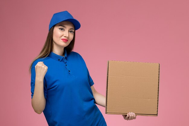 Vue de face jeune femme courrier en uniforme bleu et cape tenant la boîte de livraison de nourriture sur le mur rose clair