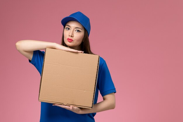 Vue de face jeune femme courrier en uniforme bleu et cape tenant la boîte de livraison de nourriture sur le mur rose clair