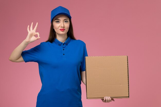 Vue de face jeune femme courrier en uniforme bleu et cape tenant la boîte de livraison de nourriture sur le mur rose