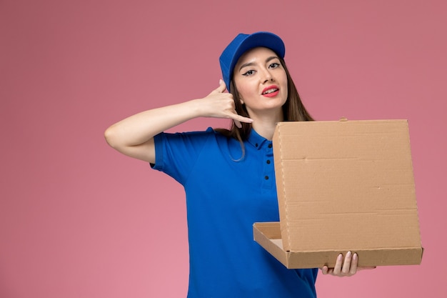 Vue de face jeune femme courrier en uniforme bleu et cape tenant la boîte de livraison de nourriture posant sur le mur rose