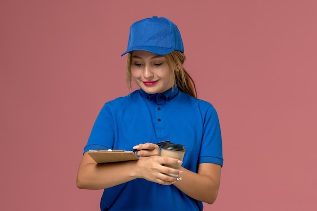 Vue de face jeune femme courrier en uniforme bleu posant tenant une tasse de café et bloc-notes, service de livraison uniforme femme travailleur