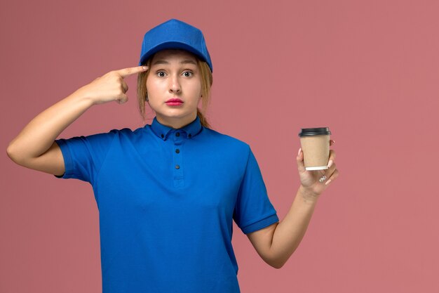 Vue de face jeune femme courrier en uniforme bleu posant tenant une tasse de café de livraison brun, service de livraison uniforme femme travailleur de l'emploi