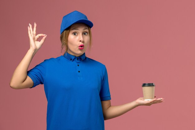 Vue de face jeune femme courrier en uniforme bleu posant tenant une tasse de café de livraison, femme de livraison uniforme de travail de service