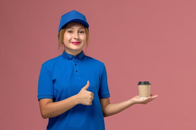 Vue de face jeune femme courrier en uniforme bleu posant tenant une tasse de café de livraison, femme de livraison uniforme de travail de service