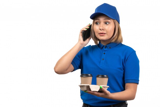 Une vue de face jeune femme courrier en uniforme bleu tenant le téléphone et les tasses à café