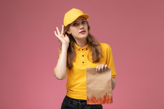 Vue de face jeune femme courrier en uniforme jaune cape jaune tenant le paquet de livraison de nourriture sur le fond rose foncé couleur de service de livraison uniforme de travail