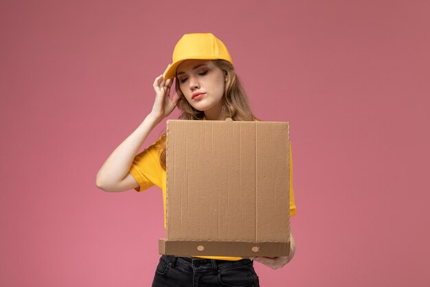 Vue de face jeune femme courrier en uniforme jaune cape jaune tenant le paquet de livraison de nourriture l'ouvrant sur fond rose foncé service de livraison uniforme travailleur féminin