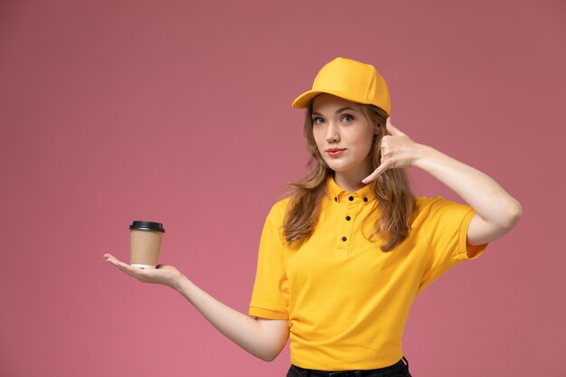 Vue de face jeune femme courrier en uniforme jaune cape jaune tenant une tasse de café sur le bureau rose foncé couleur de service de livraison uniforme