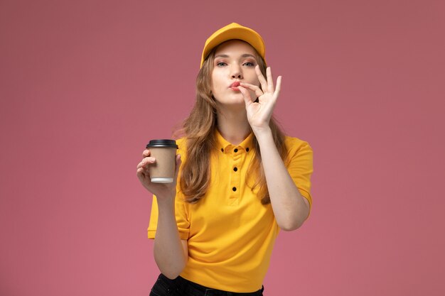 Vue de face jeune femme courrier en uniforme jaune cape jaune tenant une tasse de café en plastique sur le fond rose foncé couleur du service de livraison uniforme