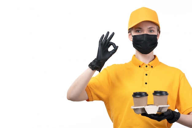 Photo gratuite une vue de face jeune femme courrier en uniforme jaune gants noirs et masque noir tenant des tasses à café montrant bien signe