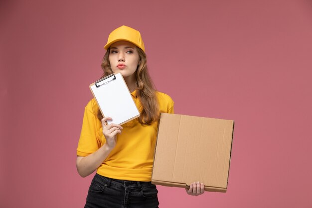 Vue de face jeune femme courrier en uniforme jaune tenant le bloc-notes et le colis de livraison de nourriture sur fond rose travail uniforme de livraison couleur service worker