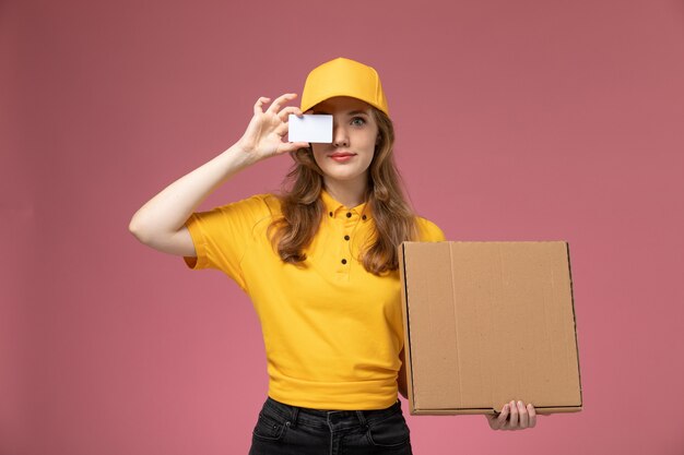 Vue de face jeune femme courrier en uniforme jaune tenant boîte brune et carte blanche sur le service de livraison uniforme de bureau rose foncé travailleur féminin