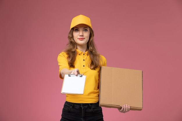 Vue de face jeune femme courrier en uniforme jaune tenant une boîte de nourriture brune et bloc-notes sur le bureau rose travailleur de service de livraison uniforme