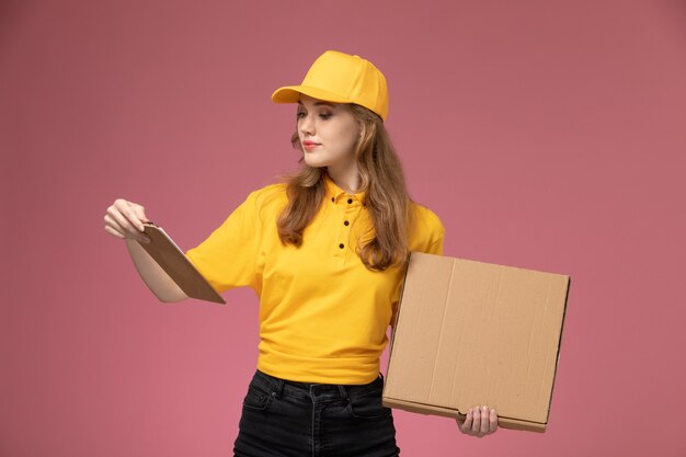 Vue de face jeune femme courrier en uniforme jaune tenant la boîte de nourriture de livraison et le bloc-notes sur le bureau rose travailleur de service de livraison uniforme