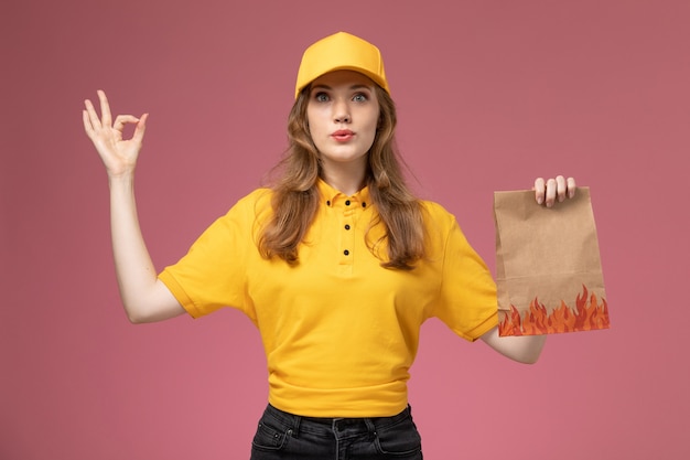 Vue de face jeune femme courrier en uniforme jaune tenant le paquet de nourriture sur le service de livraison uniforme de bureau rose foncé travailleur féminin