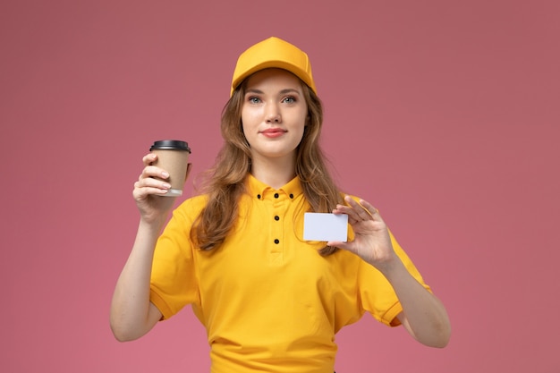 Vue de face jeune femme courrier en uniforme jaune tenant une tasse de café et une carte blanche sur le bureau rose foncé travail de livraison uniforme travailleur de service