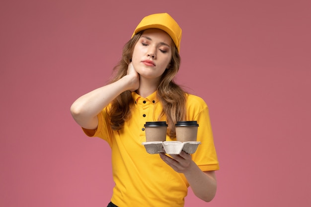 Vue de face jeune femme courrier en uniforme jaune tenant des tasses à café ayant des maux de tête sur fond rose