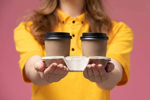 Vue De Face Jeune Femme Courrier En Uniforme Jaune Tenant Des Tasses à Café Marron En Plastique Sur Fond Rose Foncé Travail De Livraison Uniforme Couleur Service Worker