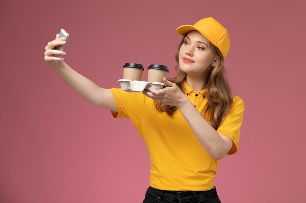 Vue de face jeune femme courrier en uniforme jaune tenant des tasses à café marron en plastique prenant selfie avec eux sur fond rose travail uniforme de livraison couleur service worker