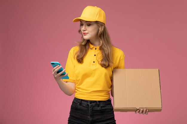 Vue de face jeune femme courrier en uniforme jaune tenant le téléphone et le paquet de nourriture sur le bureau rose foncé service de livraison uniforme travailleur féminin