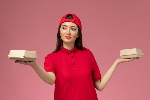 Vue de face jeune femme courrier en uniforme rouge et cape avec peu de colis alimentaires de livraison sur ses mains sur un mur rose clair