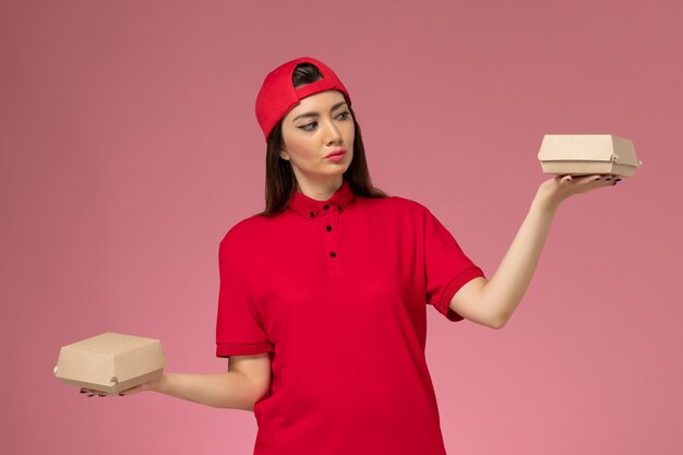 Vue de face jeune femme courrier en uniforme rouge et cape avec peu de colis alimentaires de livraison sur ses mains sur le mur rose