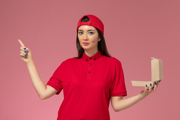 Vue de face jeune femme courrier en uniforme rouge et cape avec peu de colis de livraison de nourriture sur ses mains sur un mur rose clair